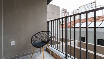 A balcony with a black wireframe chair, a tiled floor, and a view of a building with a red-tiled roof.