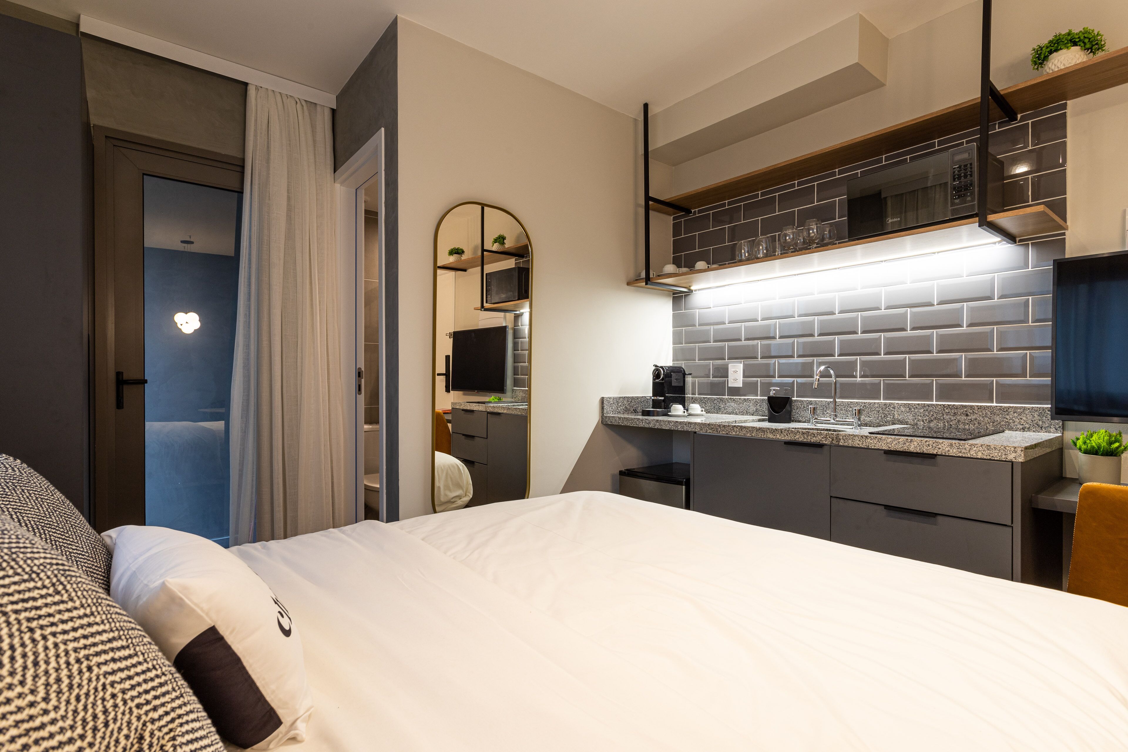 A modern hotel room with a large bed, a kitchenette with a microwave and coffee machine, and a bathroom visible through a door.