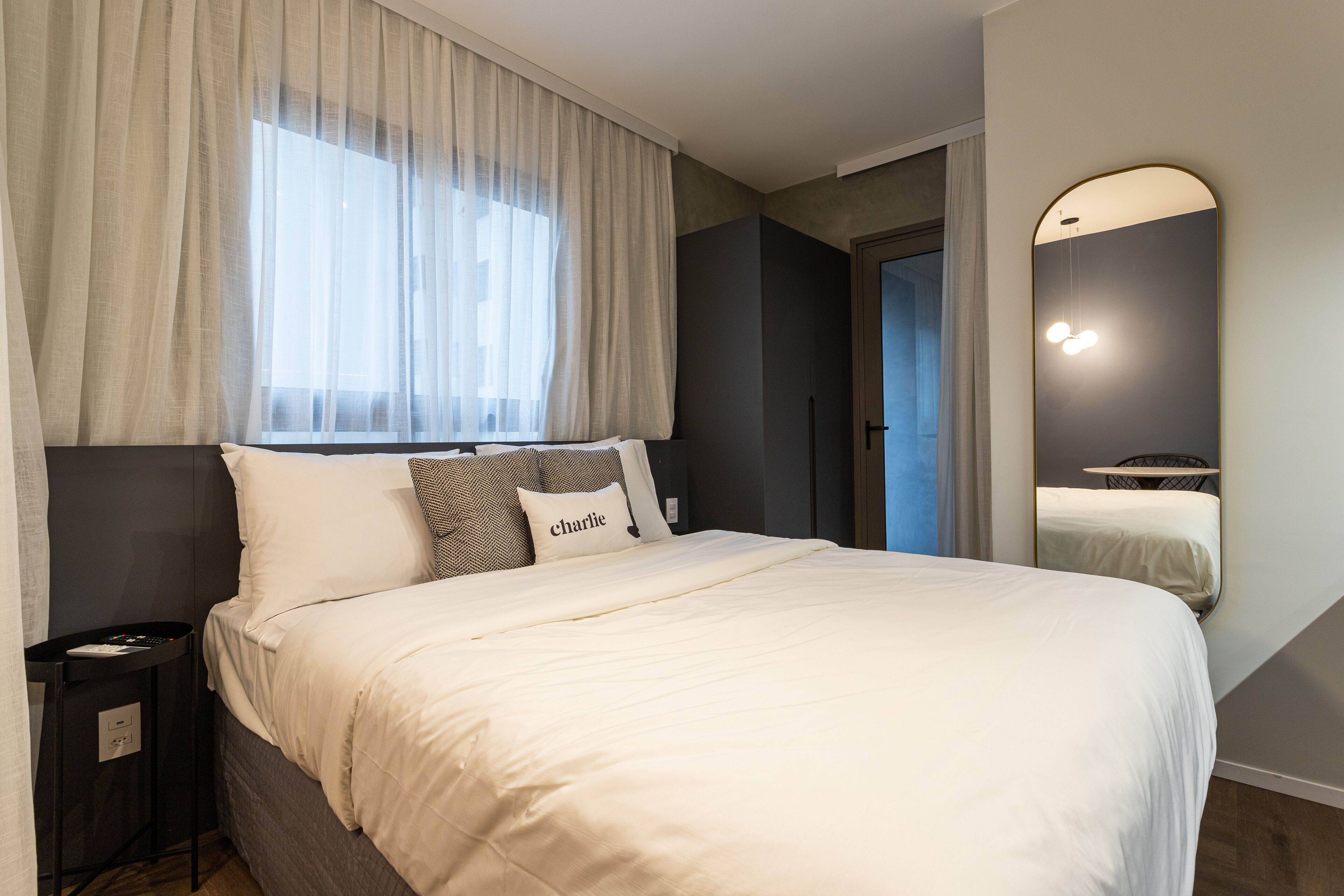 A modern bedroom with a large bed, a bedside table, a mirror, and a window with curtains.