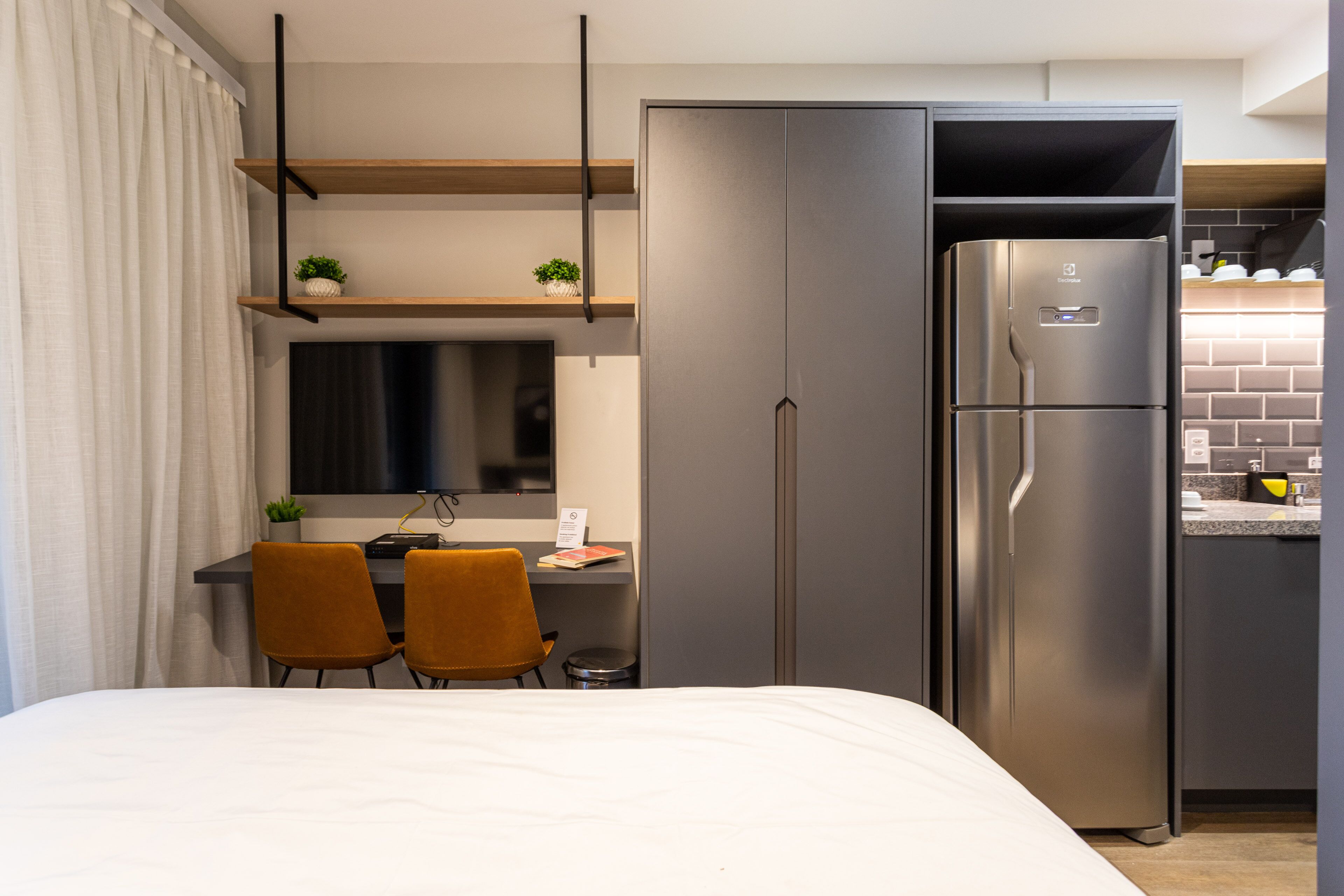 Een moderne hotelkamer met een bed, twee stoelen, een bureau, een koelkast en een kleine keukenhoek.