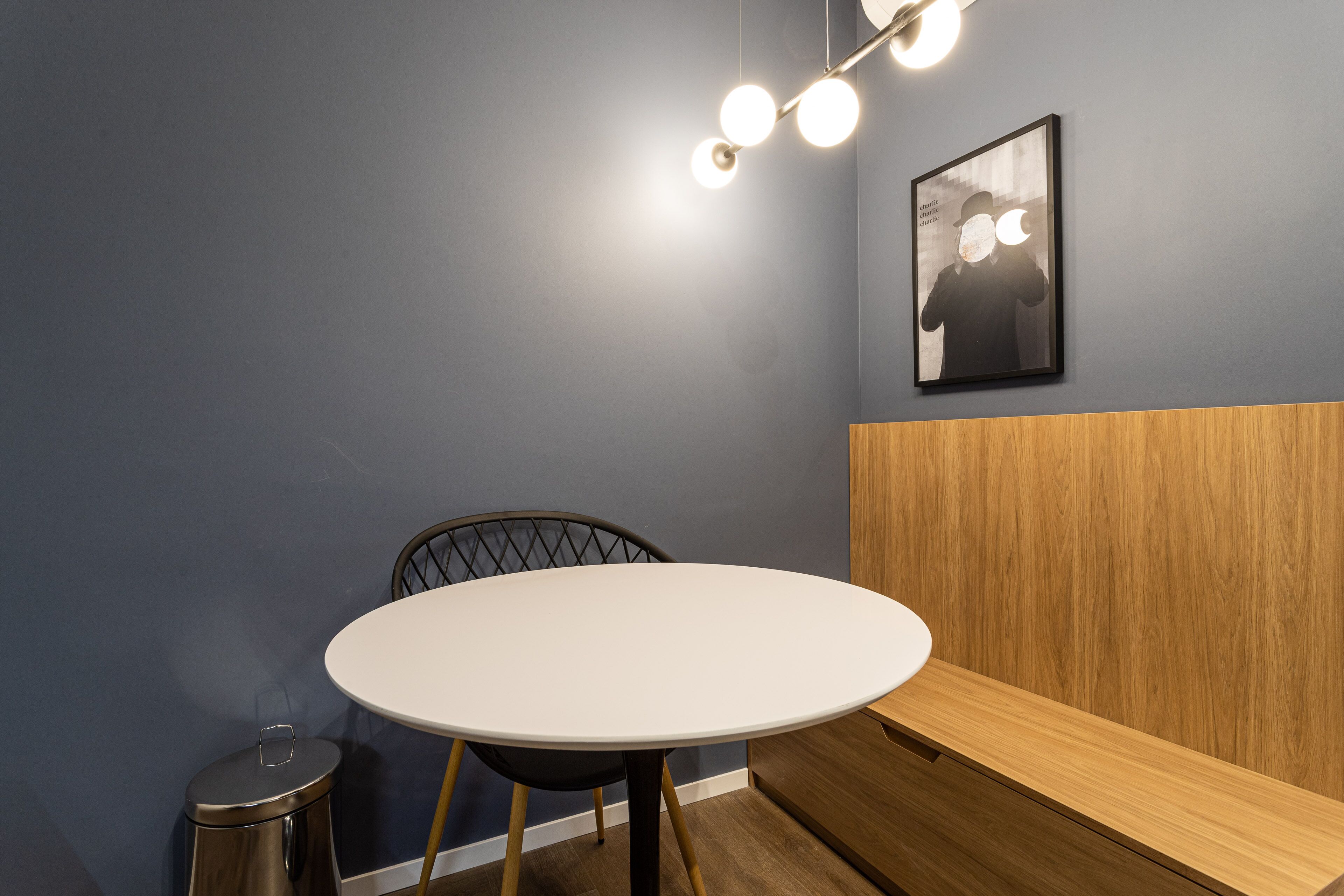 A small, round table with a white top, a black chair, a trash bin, and a wooden bench against a blue wall with a framed picture.