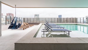 Un área de piscina en la azotea con sillones de descanso, una hamaca colgante y vistas al horizonte de la ciudad.