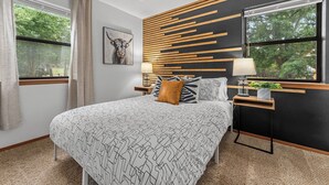 A modern bedroom with a large bed, a headboard with horizontal wooden slats, a side table with a lamp, and a window with a view of trees. - The ultimate retreat! Enjoy our hot tub, game room, and serene deer sightings. (Fredericksburg)
