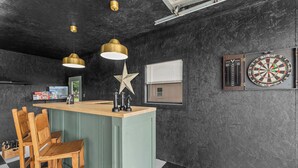 A bar area with a wooden counter, two hanging pendant lights, a star decoration, a dartboard, and a window with blinds. - The ultimate retreat! Enjoy our hot tub, game room, and serene deer sightings. (Fredericksburg)