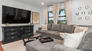 A modern living room with a large flat-screen TV, a grey sofa, a black entertainment unit, and a glass coffee table. - The ultimate retreat! Enjoy our hot tub, game room, and serene deer sightings. (Fredericksburg)