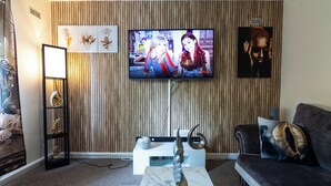 Een woonkamer met een flatscreen televisie bevestigd aan een houten wand, een zwarte bank met gekleurde kussens, een witte bijzettafel en een marmeren salontafel.