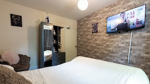 Een hotelkamer met een groot bed, een kledingkast, een spiegel, een televisie bevestigd aan de bakstenen muur, en een klein plankje met decoratieve objecten.