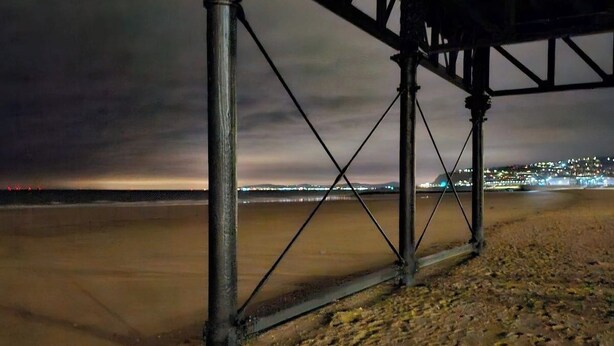 Escena nocturna en la playa con una estructura metálica, posiblemente un muelle, y luces de la ciudad a lo lejos.