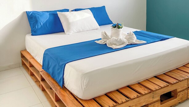 Una cama con una colcha azul y blanca, base de palets de madera y un almohadón blanco.