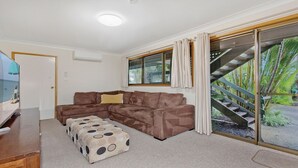 Un salon avec un canapé d’angle marron, un pouf à motifs, un téléviseur posé sur un meuble en bois et une grande fenêtre avec des rideaux.