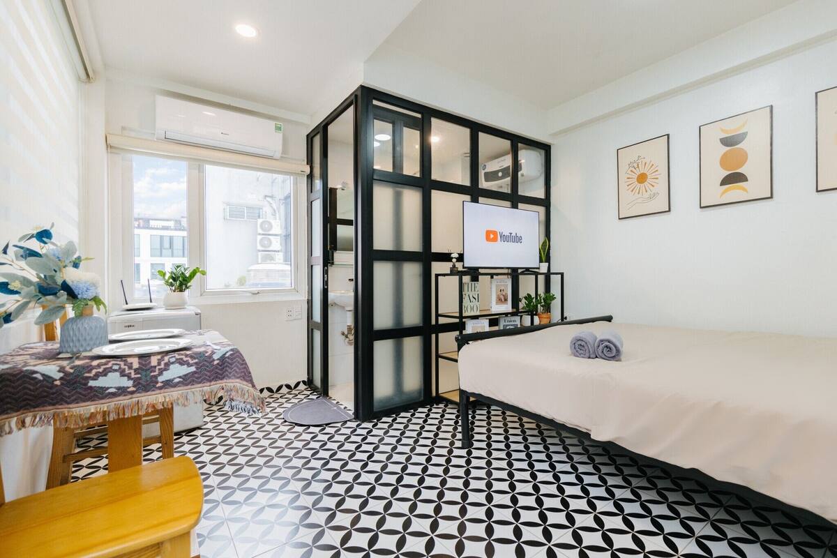 A modern room with a bed, a desk with a computer, a TV, and a window with blinds.