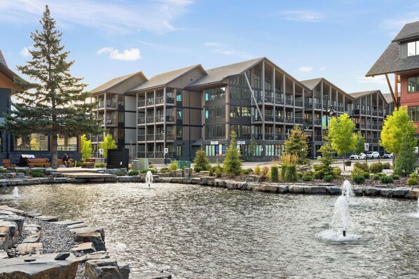 Un bâtiment moderne doté de nombreux balcons et de grandes fenêtres, situé à côté d’un élément décoratif aquatique comprenant une fontaine.