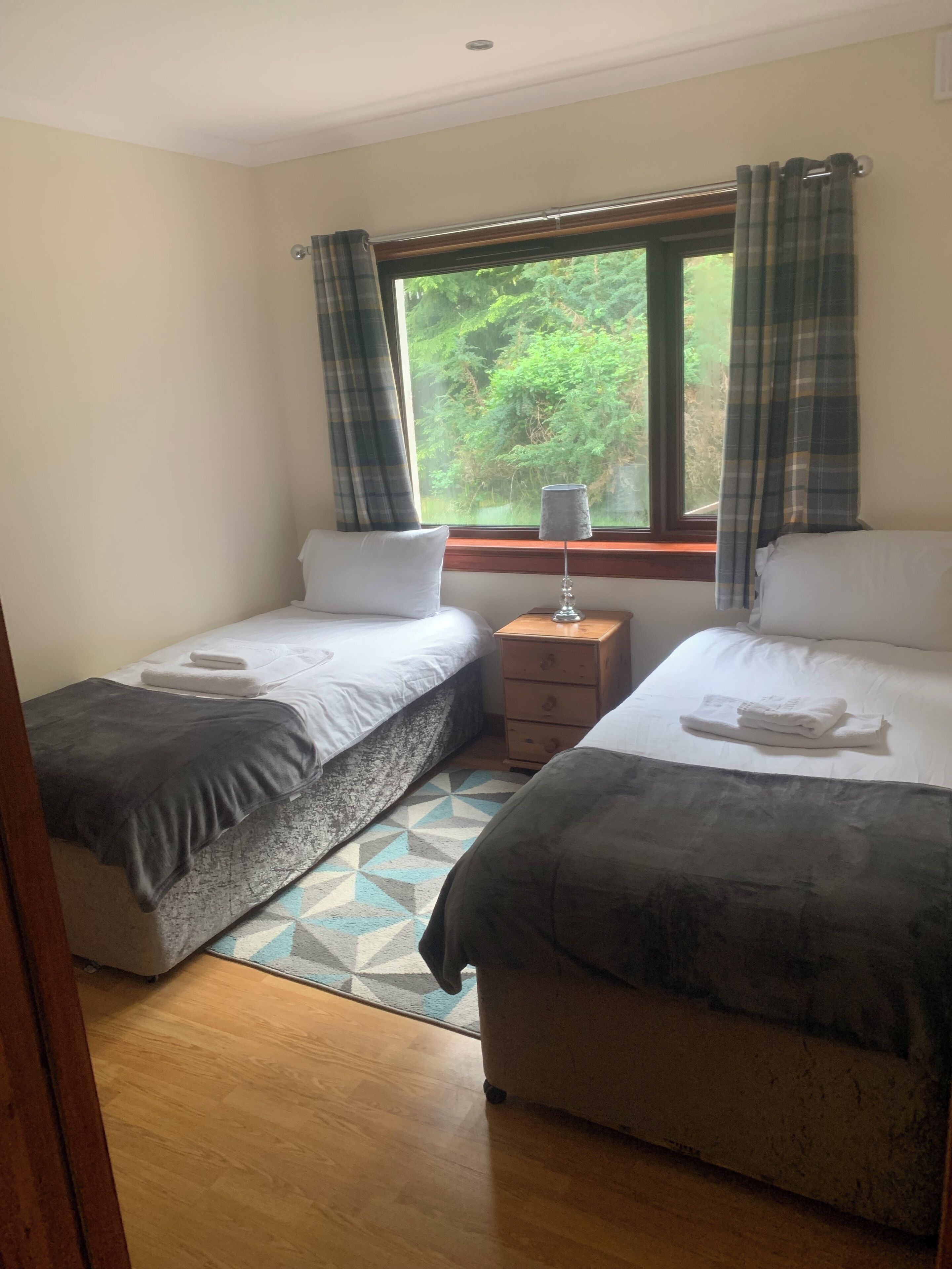 Una habitación con dos camas individuales, una mesita de noche de madera, una lámpara y una ventana con cortinas.