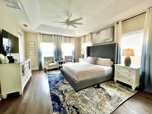A bedroom with a bed, two chairs, a nightstand, a lamp, a ceiling fan, and a large window with curtains.