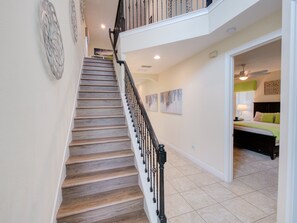 Stairs lead to a cozy upper floor with a bed and nightstand, while the lower floor features a large window and ceiling fan.