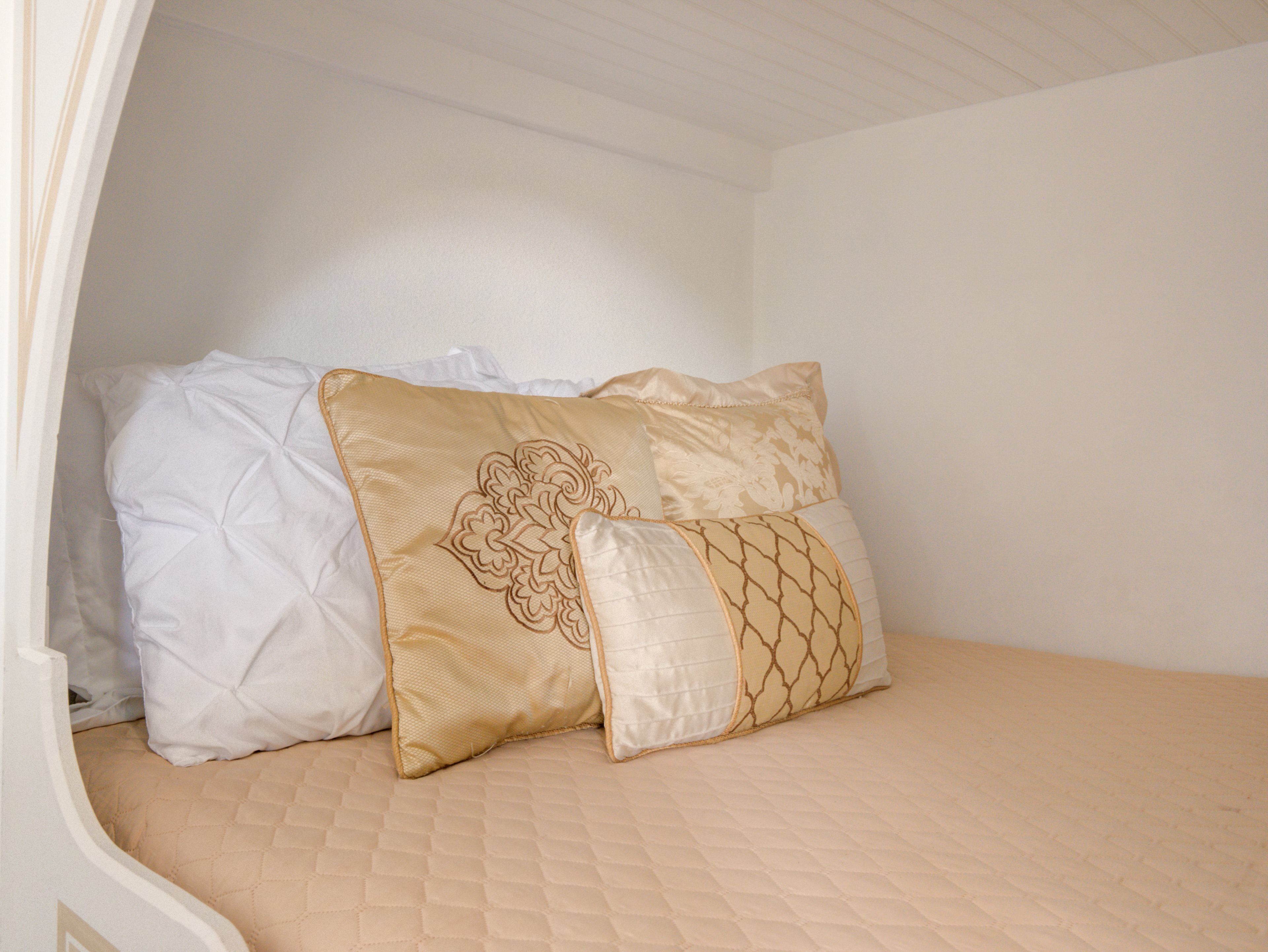 Cama com duas almofadas, uma branca e outra com padrão dourado, em quarto com paredes e teto brancos.
