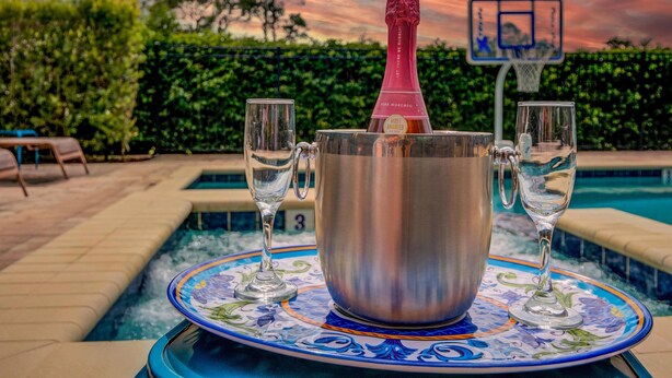Une bouteille de champagne dans un seau métallique, avec deux verres posés sur une table près d’une piscine, le tout sur fond de coucher de soleil.