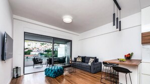 A modern living room with a sofa, ottoman, and bar stools. There is a large window offering a view of buildings and greenery. - Exclusive Apartment Lara - One Bedroom Apartment with Balcony (Dubrovnik)