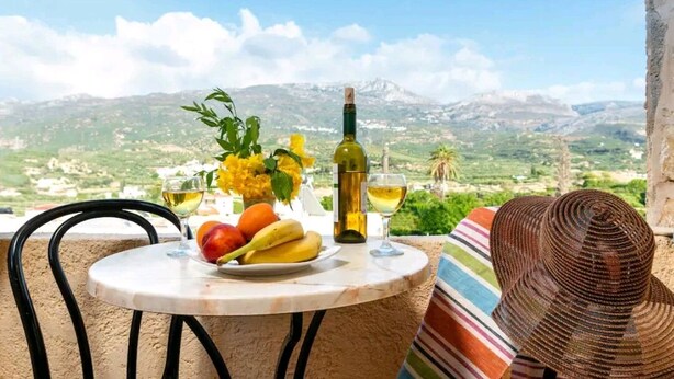 Een tafel met een fles wijn, een glas en een schaal fruit, met een bergachtig landschap op de achtergrond.