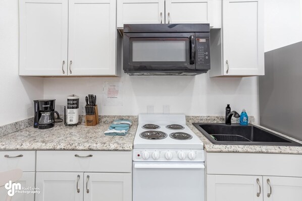 A kitchen with white cabinets, a black microwave, a white stove, a granite countertop, a coffee maker, a paper towel dispenser, and a knife block.
