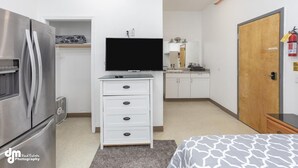 Habitación de hotel compacta con kitchenette, televisión de pantalla plana, puerta de madera y cama con colcha estampada.
