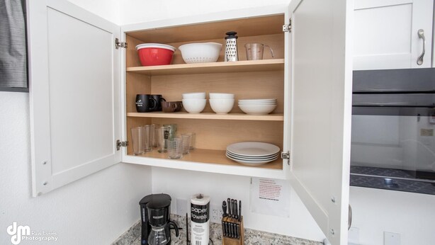 Una cocina con un armario blanco que contiene tazones, tazas y un rallador. Hay una cafetera y un juego de cuchillos sobre la encimera.
