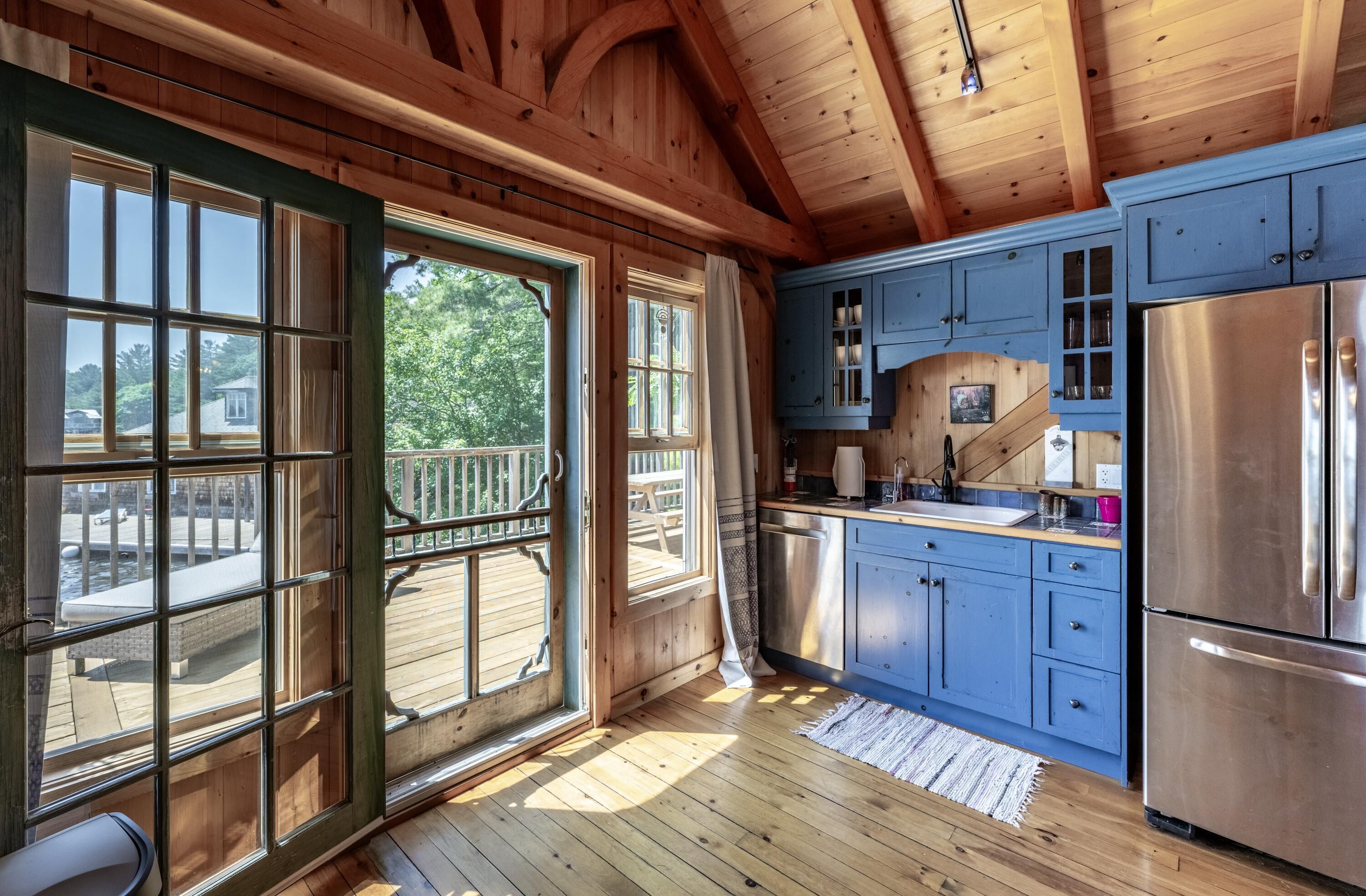 Cocina de cabaña de madera con ventanas amplias, un refrigerador de acero inoxidable y armarios azules.