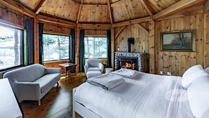 A cozy cabin interior with a bed, a sofa, a fireplace, and large windows.