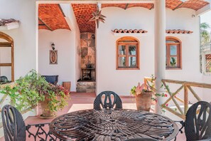 Un patio con una mesa y sillas metálicas redondas, rodeado de plantas en macetas y un muro de ladrillo.