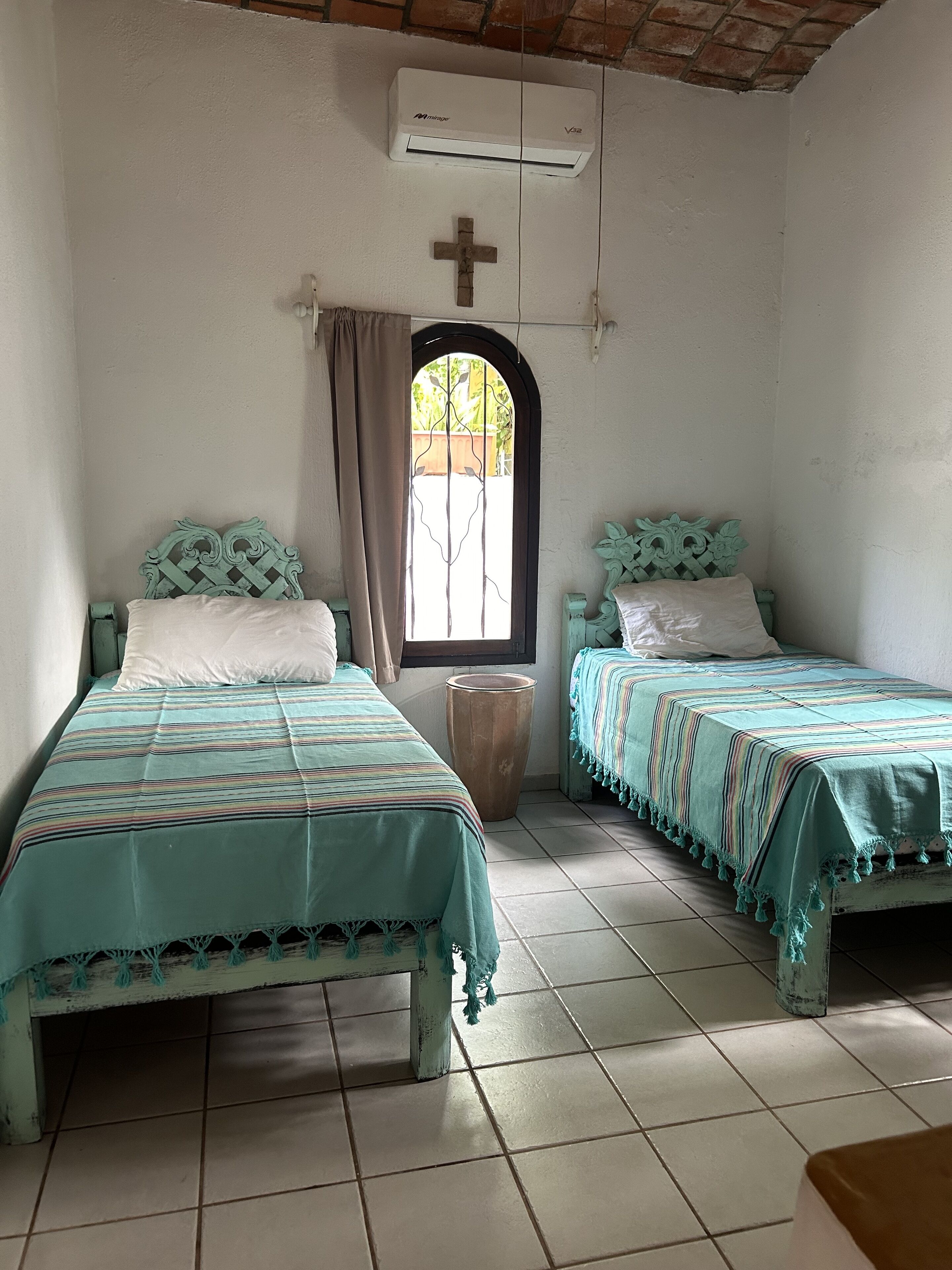 A room with two beds, a window with a cross, and an air conditioning unit.