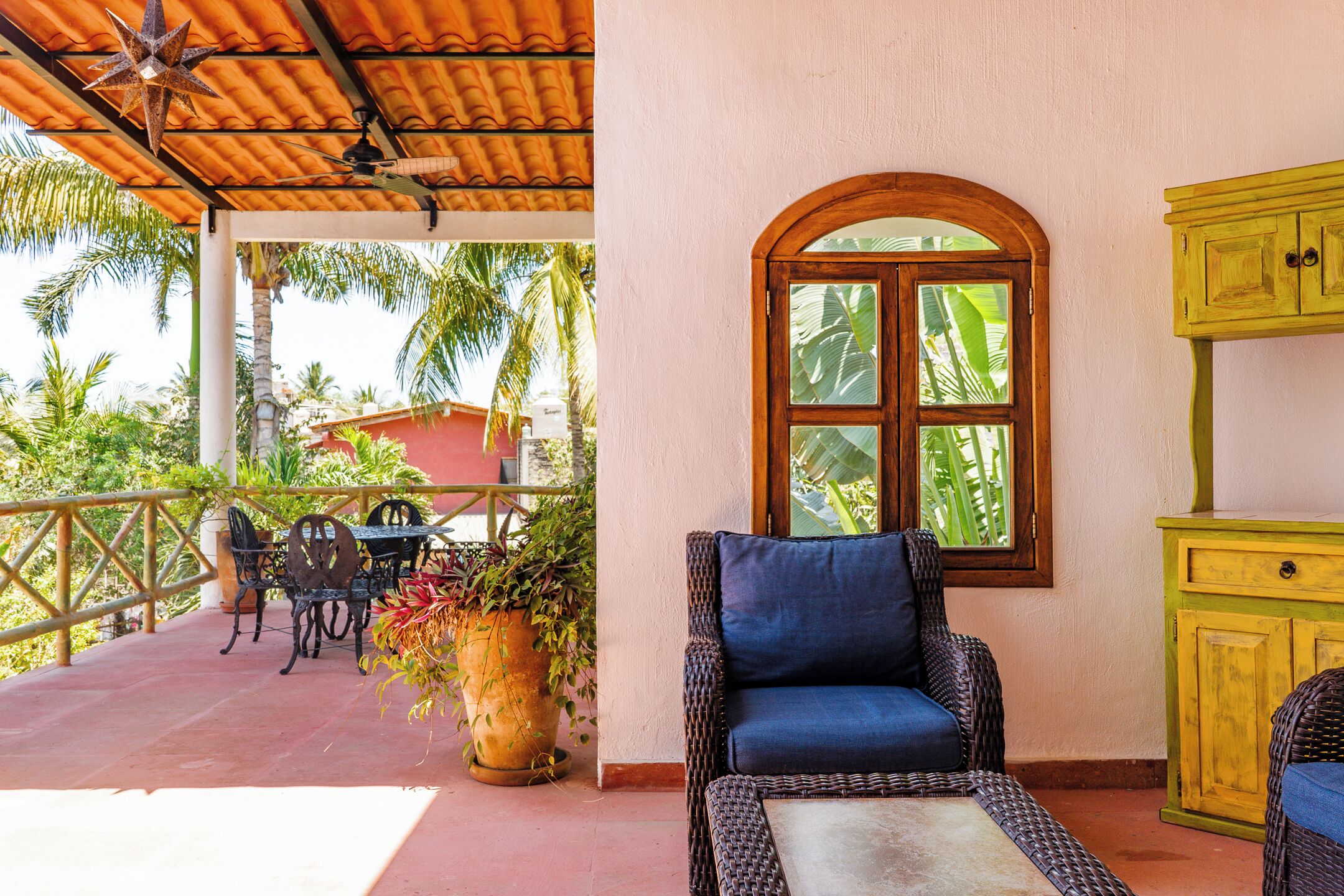 A covered outdoor seating area with wicker furniture, a potted plant, and a window with a view of palm trees.