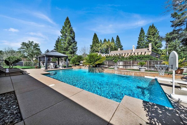 A swimming pool with a basketball hoop, lounge chairs, and a gazebo.