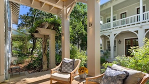 Un espace extérieur aménagé, abrité et doté de chaises en bois et d’une table, entouré d’une végétation luxuriante et bordé d’un bâtiment blanc avec un balcon.