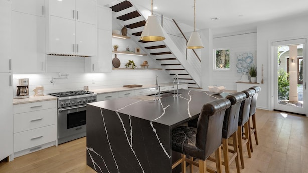 Une cuisine moderne avec des meubles blancs, un îlot central en marbre noir, des appareils électroménagers en acier inoxydable et un escalier en bois.