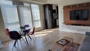 A modern living room with a grey sofa, a dining area with chairs, a TV mounted on a wooden wall, and a kitchen in the background. - Spacious Patio and Funky Vibes in Old Town! (Victoria)