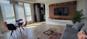 A modern living room with a grey sofa, a dining area with chairs, a TV mounted on a wooden wall, and a kitchen in the background. - Spacious Patio and Funky Vibes in Old Town! (Victoria)