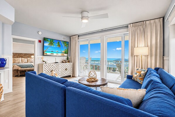 A modern living room with a large sofa, a television, and a view of the ocean through large sliding doors.
