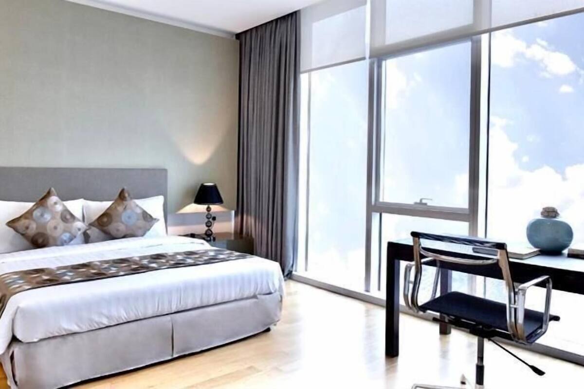 A modern hotel room with a large bed, two bedside tables with lamps, a desk with a chair, and a large window offering a view of the sky.