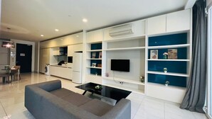 A modern living room with a sofa, a coffee table, and a flat-screen TV mounted on a white wall. There is a kitchen area with a refrigerator and a washing machine visible.