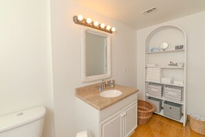 A bathroom with a white vanity, a sink, a mirror with lights, a toilet, and a shelf with towels and baskets. - Old Town Beach Cottage~ 2 blocks from Main St. and Beach Blvd. (Bay St. Louis)