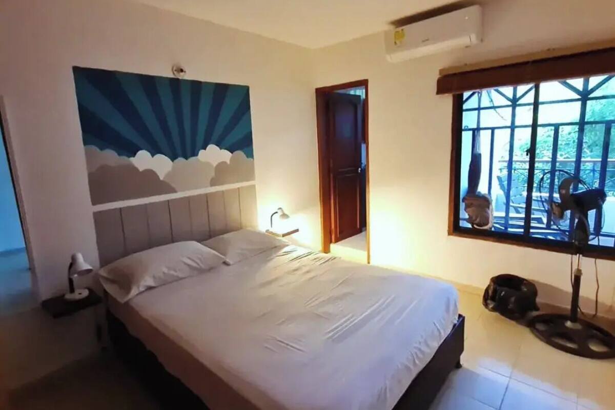 A bedroom with a bed, a nightstand, a wall mural, a window with a view, and a ceiling fan.