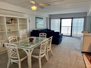 A room with a glass-top dining table, white wicker chairs, a dark blue sofa, and a ceiling fan. - High Point South 4C - 2 Bedroom Oceanfront condo, Oceanfront POOL (Ocean City)
