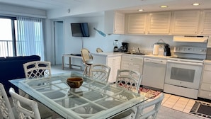 A compact kitchen with white cabinetry, a dining area with a glass table, and a TV mounted on the wall.