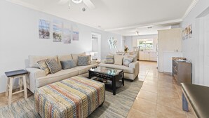 A spacious living room with a sofa, ottoman, and coffee table, a dining area with chairs, and a kitchen in the background.