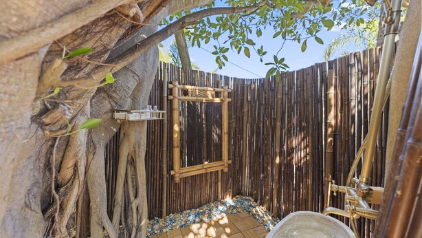 A bamboo-walled outdoor shower area with a wooden floor, surrounded by natural elements like tree roots and foliage.