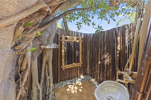Un espace de douche extérieur entouré de murs en bambou et doté d’un plancher en bois, agrémenté d’éléments naturels tels que des racines d’arbres et du feuillage.