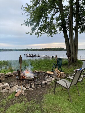 A lakeside campfire with chairs and a table, surrounded by trees and a view of boats on the water. - Come to The Lake House Loft for a relaxing visit!
 (Hammond)