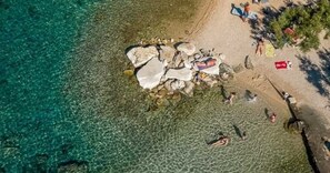 En vy över en strand sedd från luften, med klart vatten, klippor och människor.