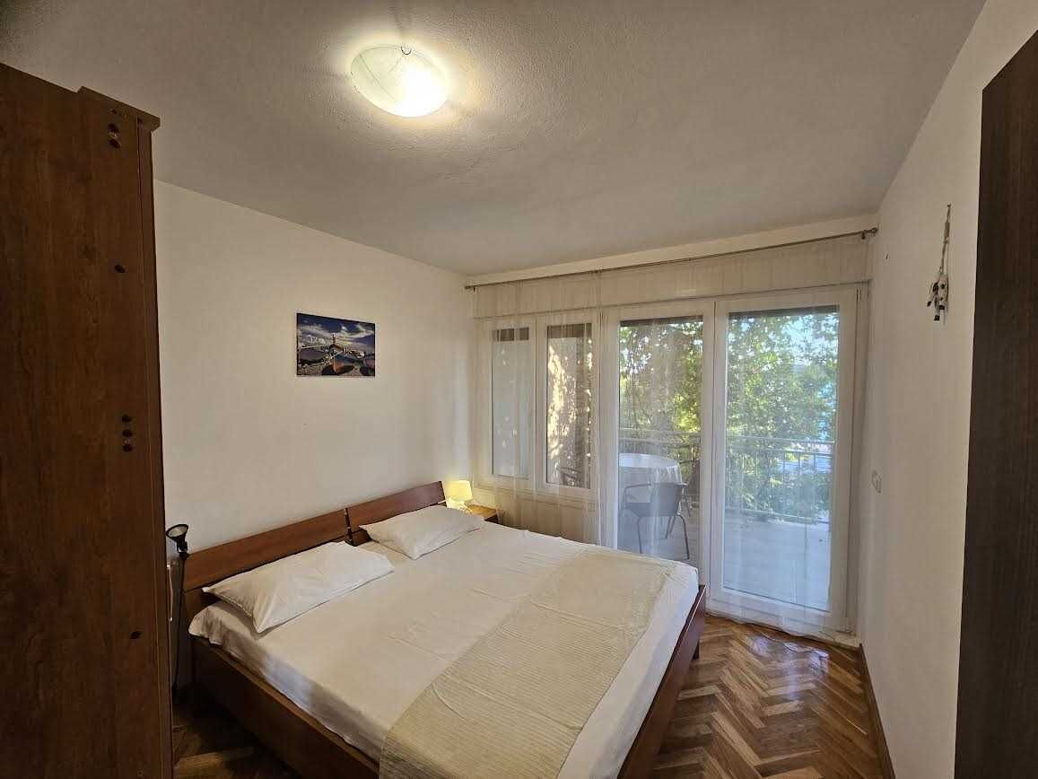 A bedroom with a large bed, a wooden wardrobe, a ceiling light, a window with curtains, and a small table with a lamp.