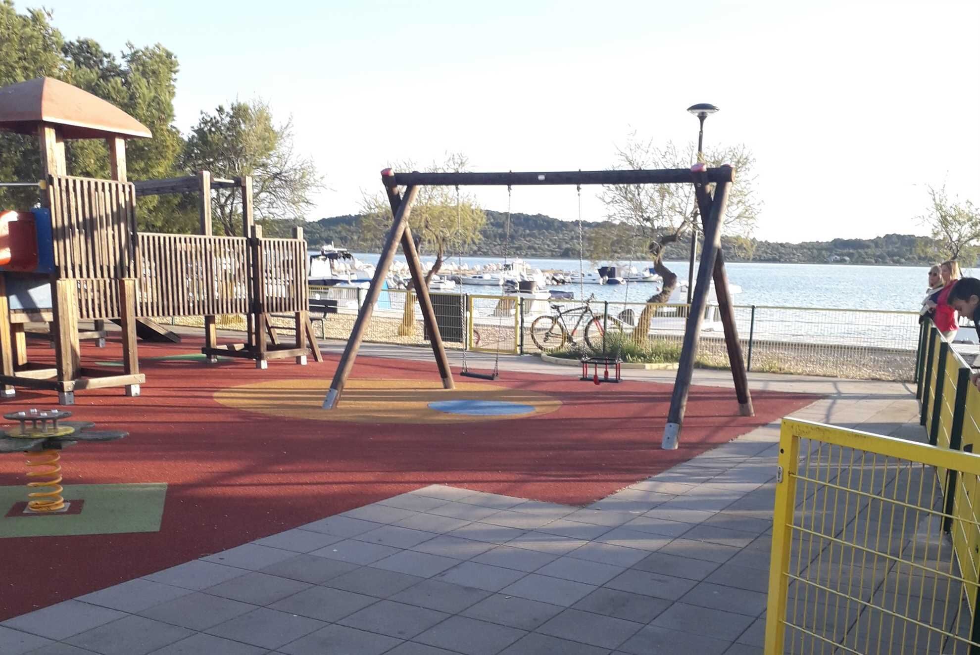 A playground with swings and a slide, near a waterfront with boats and a hilly landscape in the background.